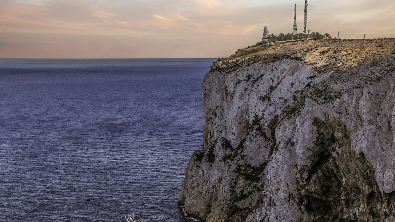 An image capturing a scenic view at dusk, featuring a white lighthouse perched on the edge of a steep, rocky cliff. Below, the calm sea extends to the horizon